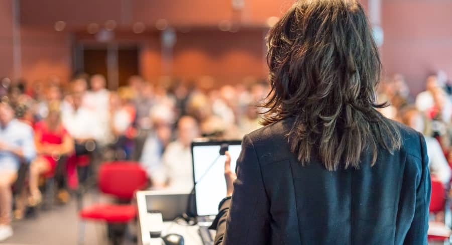 the back of a business woman behind a podium as she gives a presentation to a full conference room audience